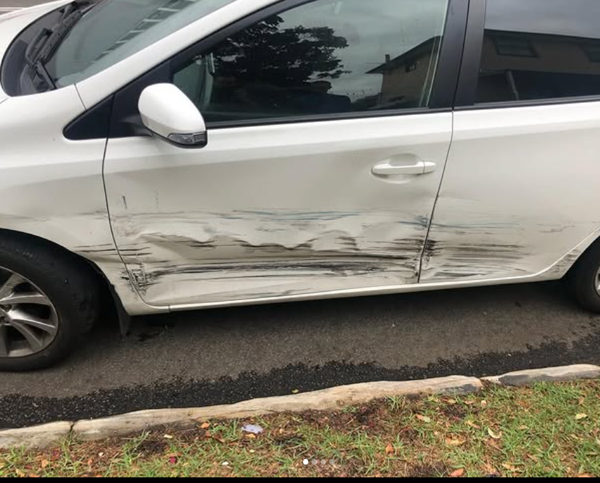 Same white hatchback before repair, with deep side impact damage along the doors